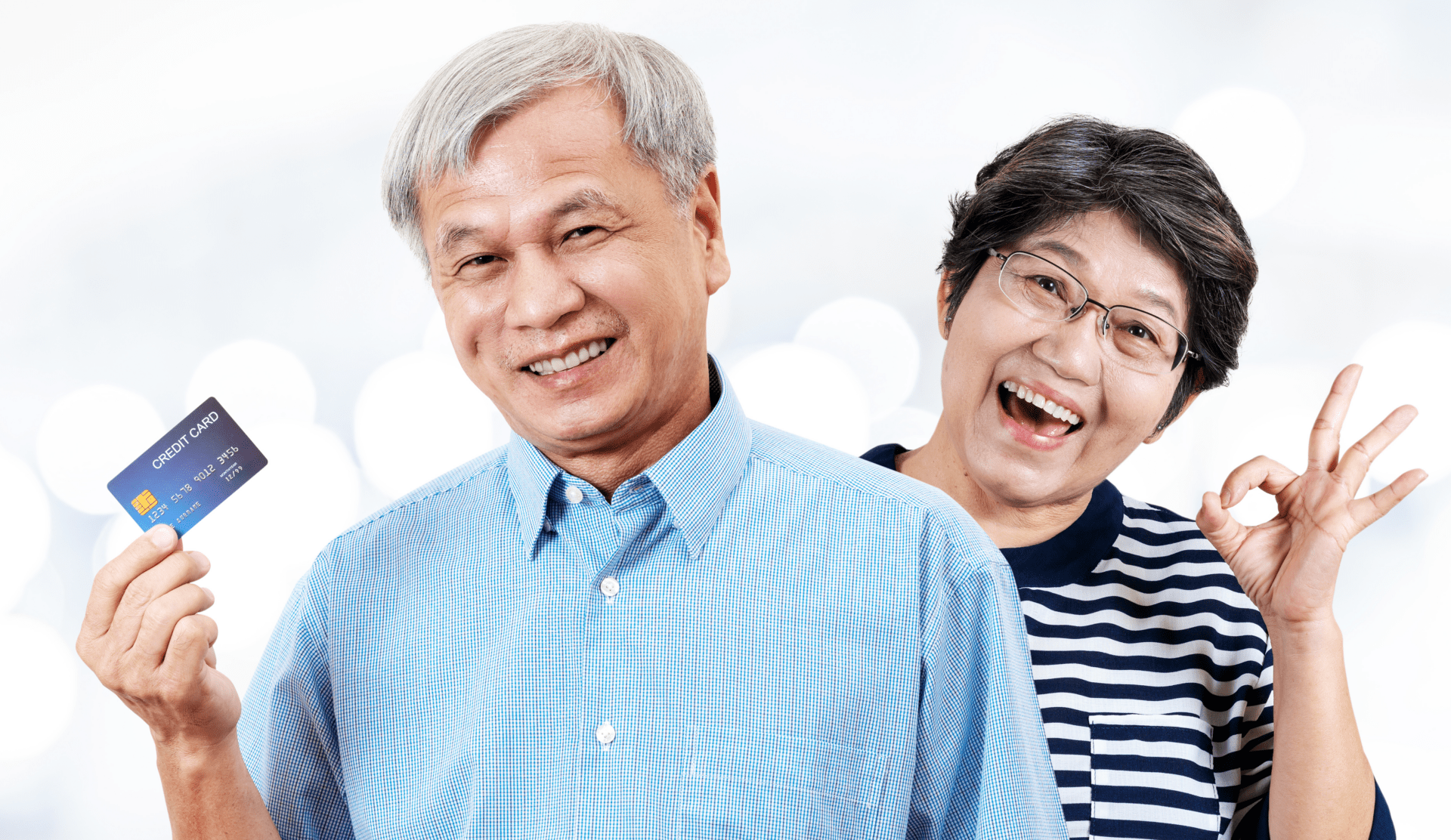 Mature Asian couple in front of white background. Man holding credit card. Woman making "OK" symbol with fingers.