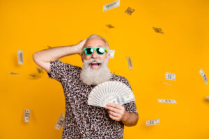 Mature Man with long beard and mustache, wearing leopard t-shirt and green sunglasses, holds fan of $100 bills. Money flying around him, against yellow background.