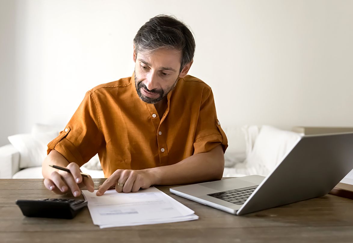 A serious and thoughtful middle aged man sitting at home desk calculating finances with paper, a calculator, and a laptop