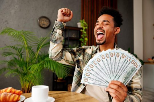 A young man smiling while sitting down in a cafe holding a fan of $100 bills in one hand with his other arm pumped in the air